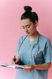 Portrait of a young nurse in scrubs writing medical notes on a clipboard against a pink background.