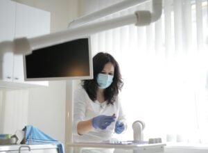 Serious female dentist in medical mask and latex gloves wearing white uniform preparing instruments for treatment in light modern dental room in clinic